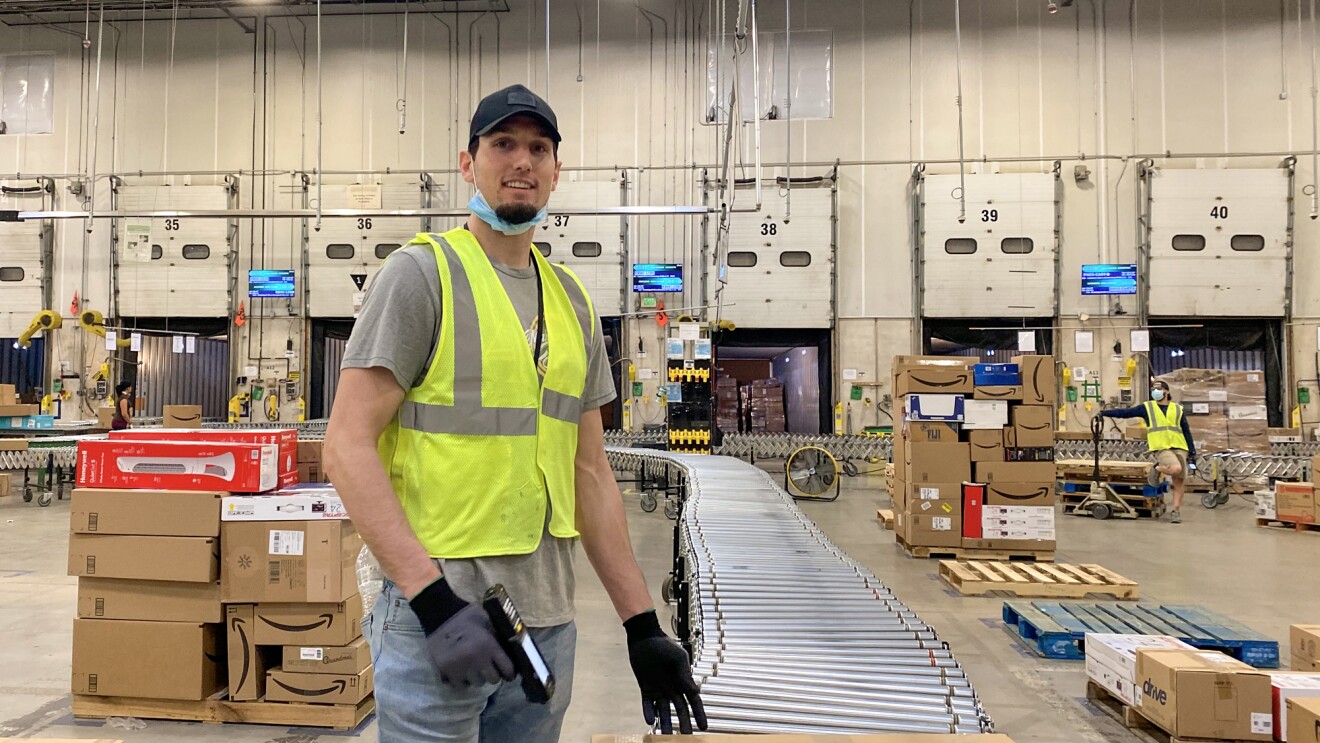 A man wearing a safety vest and a protective mask stands next to a stack of boxes holding a barcode scanner.