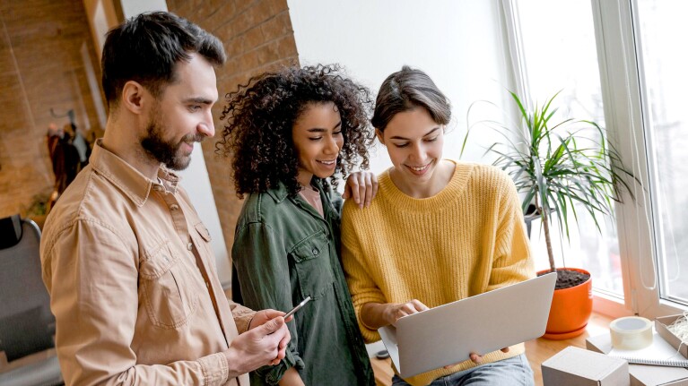 Three people stand looking excitedly at a laptop that one person is holding