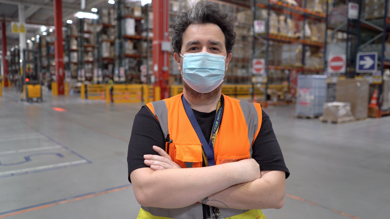 Uomo ripreso a mezzo busto in un magazzino Amazon. Indossa un giubbino catarinfrangente arancione e giallo, ha le braccia conserte. Indossa una mascherina chirurgica sul volto, ha i capelli neri con un ciuffo bianco. Alle sue spalle si intravedono gli scaffali di un magazzino Amazon. 
