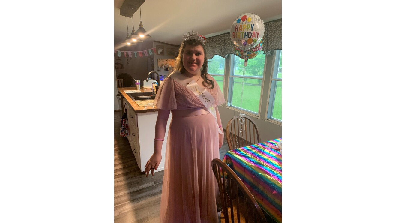 An image of a young woman standing in her kitchen smiling for a photo while wearing a flowy, pink dress, a tiara, and a sash that says "Sweet 16."