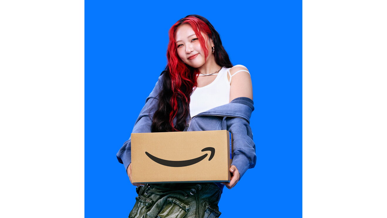 黒髪の一部を赤く染めた若い女性が、両手でAmazonロゴ入りダンボール箱を持って前に差し出している。