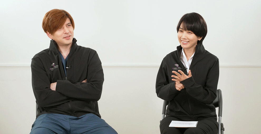 白い壁の室内で椅子に座った2人の人物が笑顔で談笑している。右側の女性は、膝の上に白い紙の束をのせている。左側の男性は腕組みをしている。