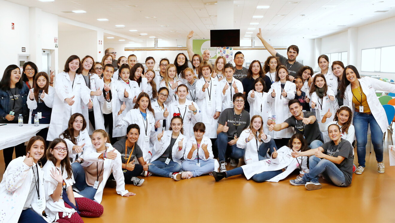 Foto de familia de la jornada  g4g @work con Amazon. Las 40 participantes, de entre 10 y 13 años, los responsables de la jornada y los voluntarios de Amazon aparecen en una foto conjunta. Algunos de ellos están sentados, otros de pie y algunos saludando a cámara. Todas las participantes lucen bata blanca. 