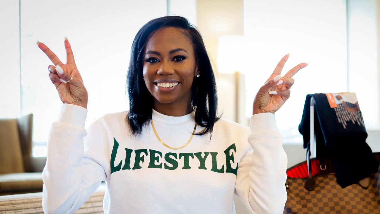 An mage of a woman smiling for a photo while making two peace signs with her hands. She is wearing a white sweatshirt that says "LIFESTYLE" on it.