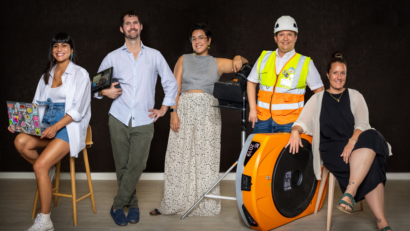 Es una foto de cinco trabajadores de Amazon posando en un tono informal. Hay tres mujeres y dos hombres. Las dos mujeres que están a la izquierda y a la derecha están sentadas y el resto de pie. Empezando de izquierda a derecha. Una mujer morena con el pelo largo, sentada en un taburete con su ordenador. Lleva una camisa blanca  con un jersey blanco debajo y unos pantalones cortos. Lleva una deportivas. El hombre de sui izquierda lleva el pelo corto y barba. Agarra una tablet. Lleva camisa azul por encima de un pantalón verde. La mujer que está en el centro está apoyada en un foto de cine de color negro. Lleva gafas, el pelo recogido, un jersey de color gris y una falda larga blanca con todos grises y negros. El hombre de su izquierda lleva casco y chaleco y está apoyado encima de un robot de Amazon. Debajo del chaleco lleva una camiseta blanca y unos jeans. La última mujer de la foto está sentada en una silla. Lleva el pelo recogida, apoya su brazo derecho en el robot. Lleva un vestido negro largo y por encima una chaqueta de punto. El fondo de la foto es negro.
