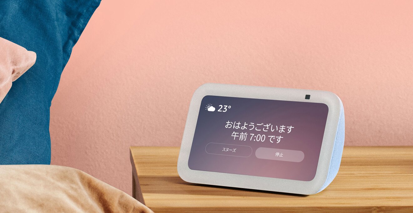 パステルピンクの壁を背景に、ベッドサイドの木製チェストに白いEcho Show端末が設置されている。側面は淡い水色。紫色のグラデーションの画面に「おはようございます 午前7:00です」の表示。