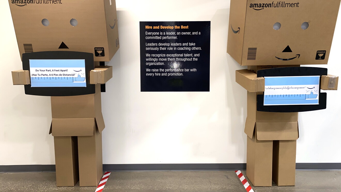 Dwie „pudełkowate osoby” wykonane z tektury stoją w centrum logistycznym Amazon. oba są ustawione w odległości 6 stóp od siebie i posiadają znaki z napisem „Wykonuj swoją część, w odległości 6 stóp od siebie”