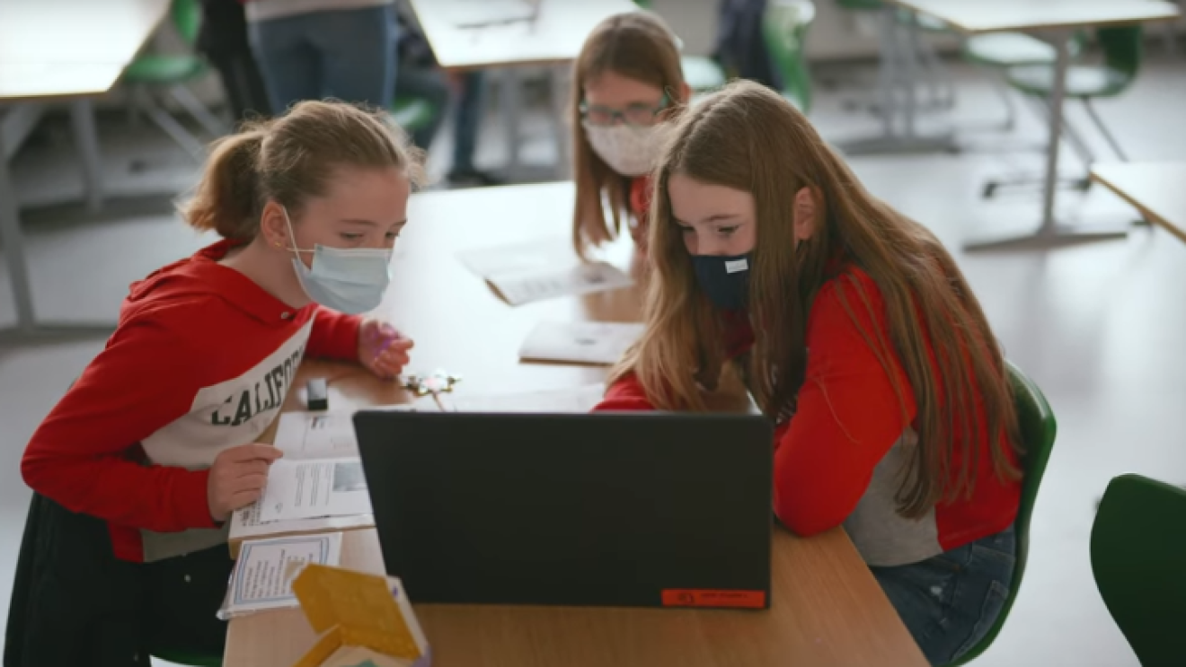 Drei Mädchen mit Masken sitzen in einem Klassenzimmer an einem Tisch zusammen und schauen in einen Laptop.