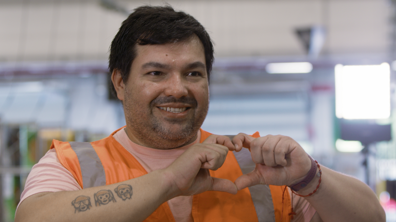 Primo piano di dipendente Amazon che sorride verso la camera. Con le mani mima la forma del cuore e sull'avambraccio ha un tatuaggio con 3 scimmiette