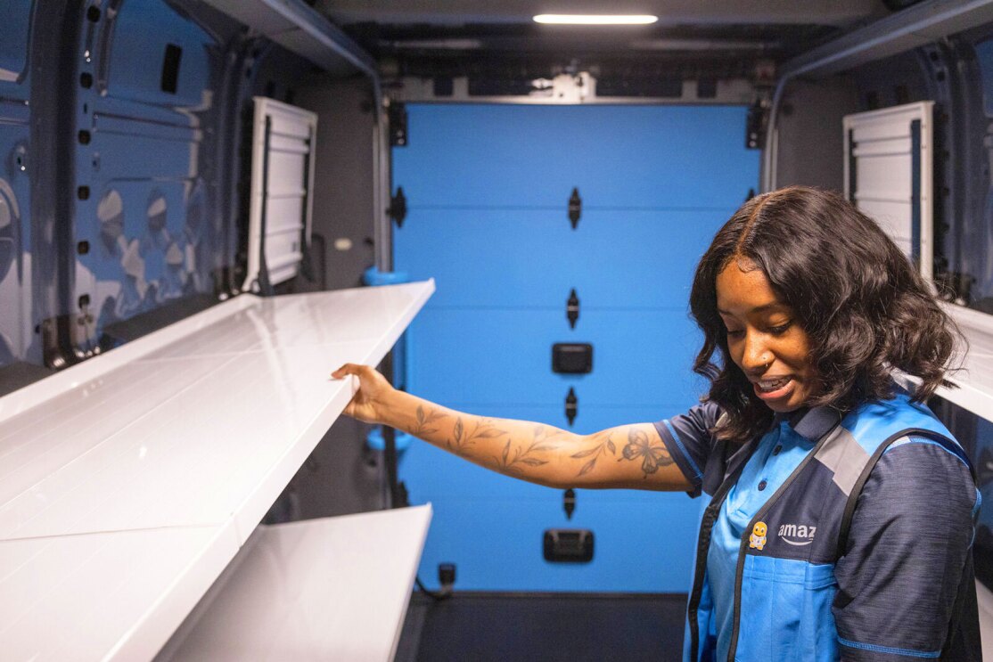 A photo of a delivery driver standing inside the storage space of an Amazon delivery van from Rivian, holding a shelf.