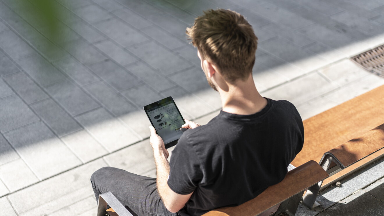 Ein junger Mann sitzt auf der Parkbank und hat ein Tablet in der Hand. Er bestellt von unterwegs bei Amazon.