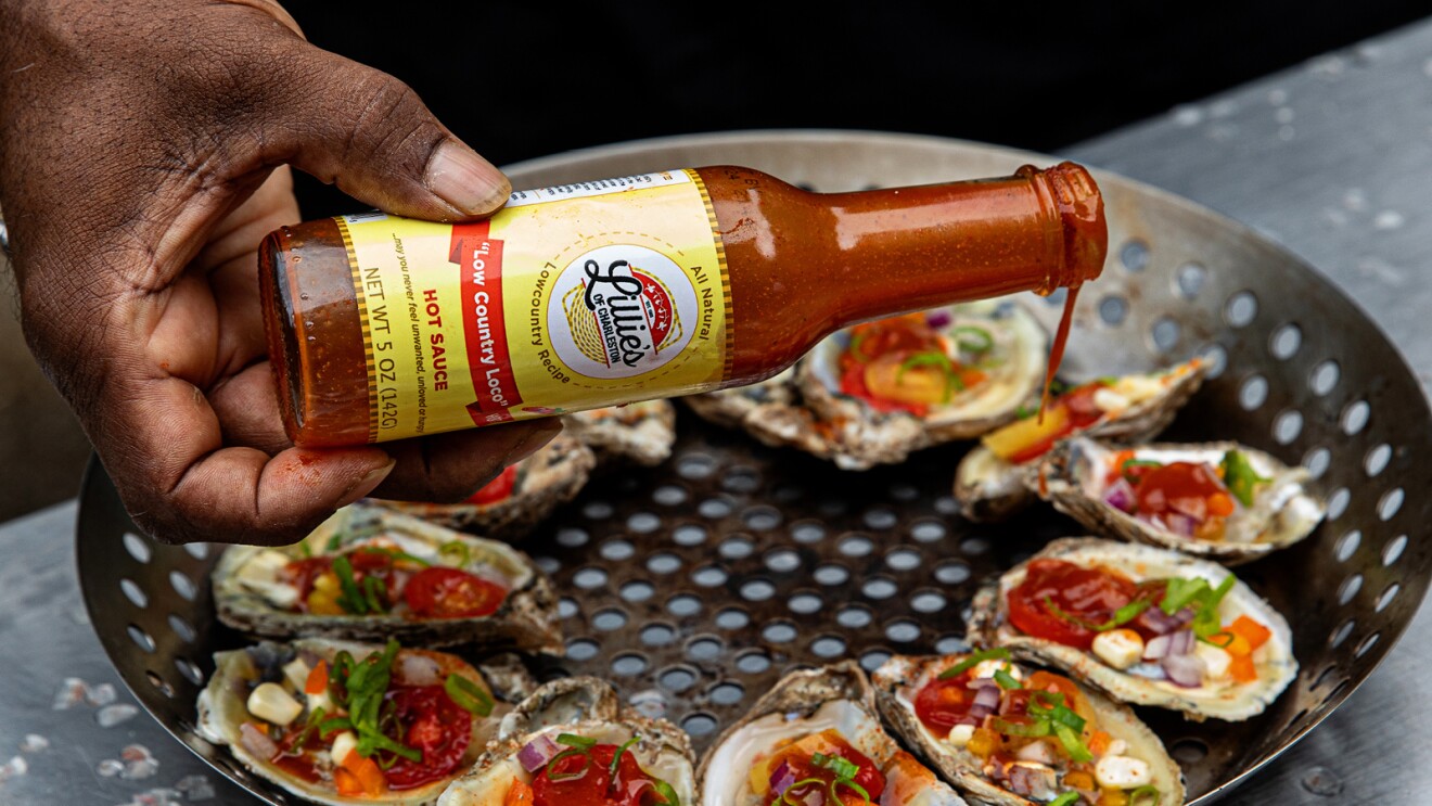 A photo of Lillie's of Charleston hot sauce being poured over barbeque oysters.
