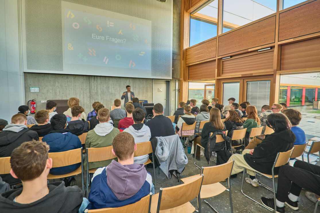 Man sieht Schüler:innen von hinten, die vor einer großen Leinwand in einem Konferenzraum sitzen. Vorne steht Amazon-Deutschlandchef Rocco Bräuniger, der vorträgt. 