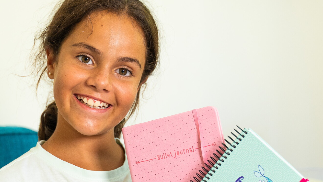  Martina tiene 11 años y es la segunda hija de la influencer Gloria Santamaría. Está sentada en un sillón azul  con dos cojines blancos con hojas verdes y negras. Mira a cámara sonriendo, en la mano derecha lleva un estuche de color rojo con corazones blanco y en la mano izquiera una agenda rosa y una libreta que pone  "hay muchos peces en el mar pero yo soy una sirena 2020". 