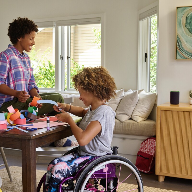 A mother sits on a dining room table as she and her daughter laugh, while making a paper chain. Behind them, Amazon Echo is on a console table. 