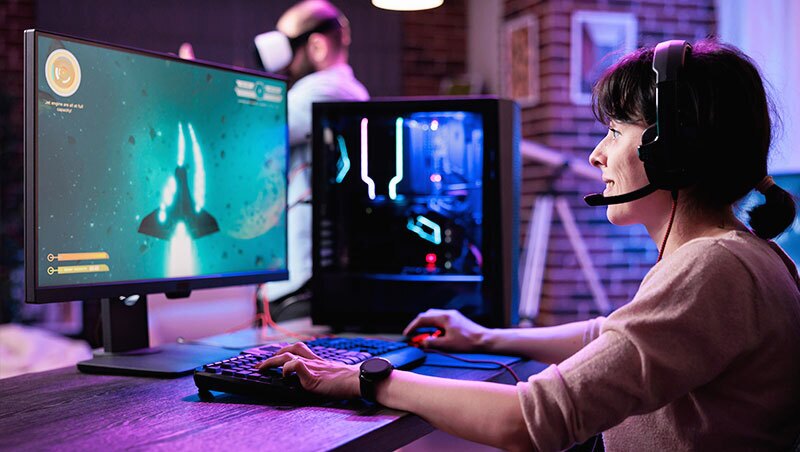 Woman sits at computer with gaming headset on playing a game on Twitch. 