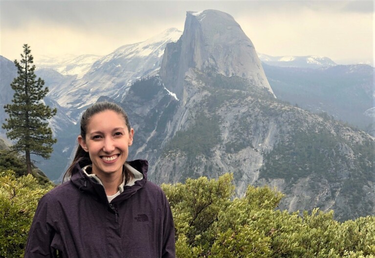 Caitlin smiles while standing outside. There is a mountain range in the near background.