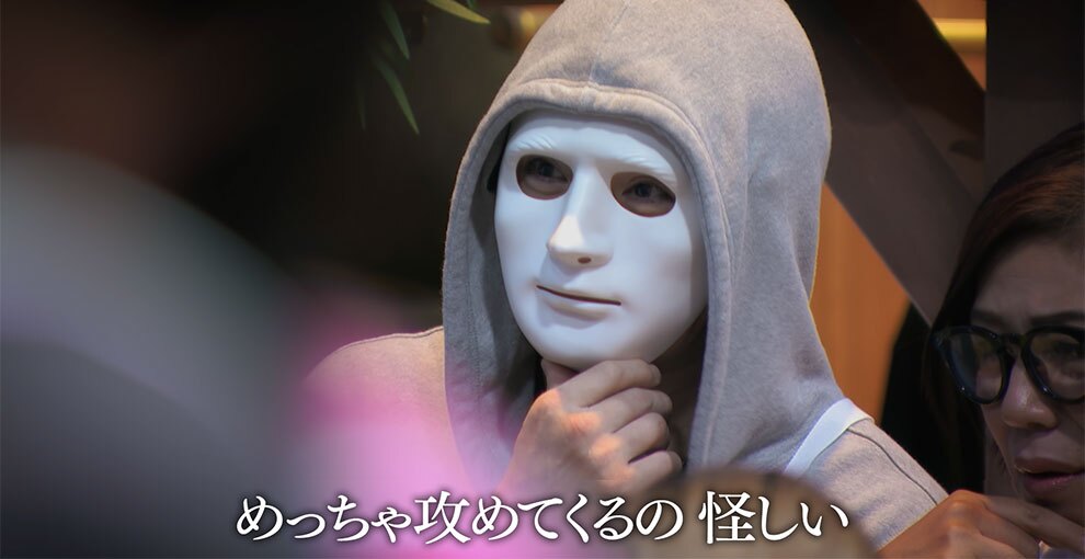 グレーのパーカー着用、フードをかぶって白い仮面をつけた人物のクローズアップ。顎に手を当てて考えるポーズ。画面下部に「めっちゃ攻めてくるの 怪しい」というテロップが表示されている。