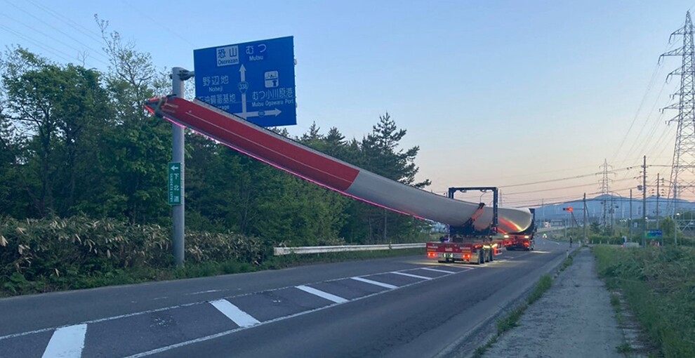 日本の自動車道で、風力発電用風車の巨大な羽を特殊車両が赤信号で停止している。羽の色は赤と白。道路上にはこの輸送車両しかない。「恐山」や「むつ」、「野辺地」などの地名が書かれた道路標識が左側に立っている。