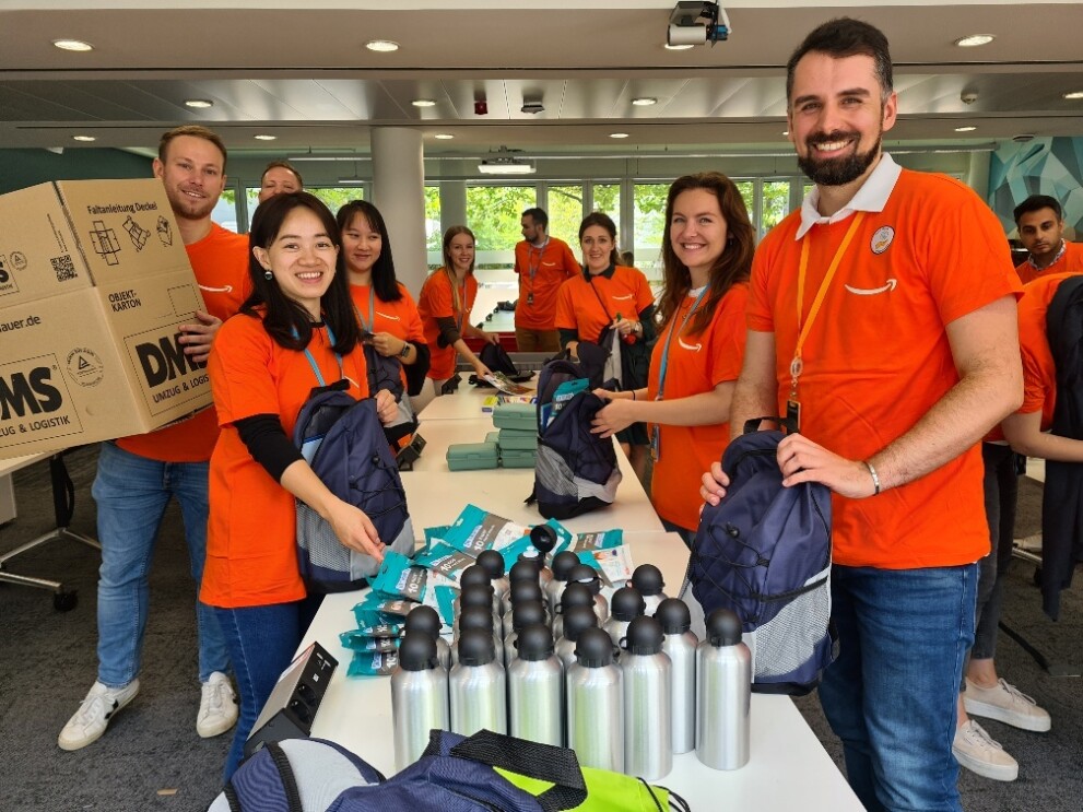Amazon Mitarbeiter:innen stehen an einem Packtisch und packen im Rahmen der Back to school Aktion von Amazon Schulutensilien für sozial beanchteiligte Kinder.