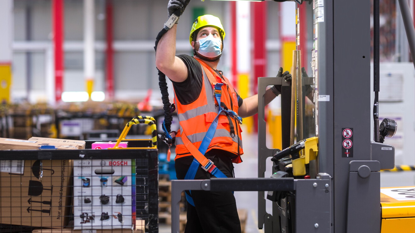 Un empleado de Amazon que se beneficia de la tecnología de arnés de seguridad en altura (en un recogepedidos o un camión industrial motorizado). En un toro hay un empleado de Amazon enganchando su arnés a la parte superior de este. Con la otra mano se sujeta a la barandilla del toro que está alzada. El toro es de color gris y amarillo y el empleado va con unos pantalones negros y una camiseta negra también de manga corta, un chaleco naranja, el arnés, una mascarilla quirúrgica y un casco de color amarillo. El toro tiene un palet agarrado con cajas blancas y maroones. De fondo se ve más material en contenedores. 
