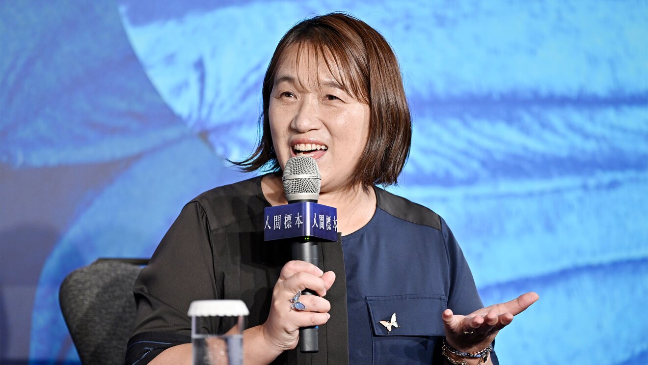 水色と紺色の壁を背景に、マイクを持って話す女性(湊かなえさん)。黒と紺が半々の服を着用し、胸ポケットに蝶の形のピン