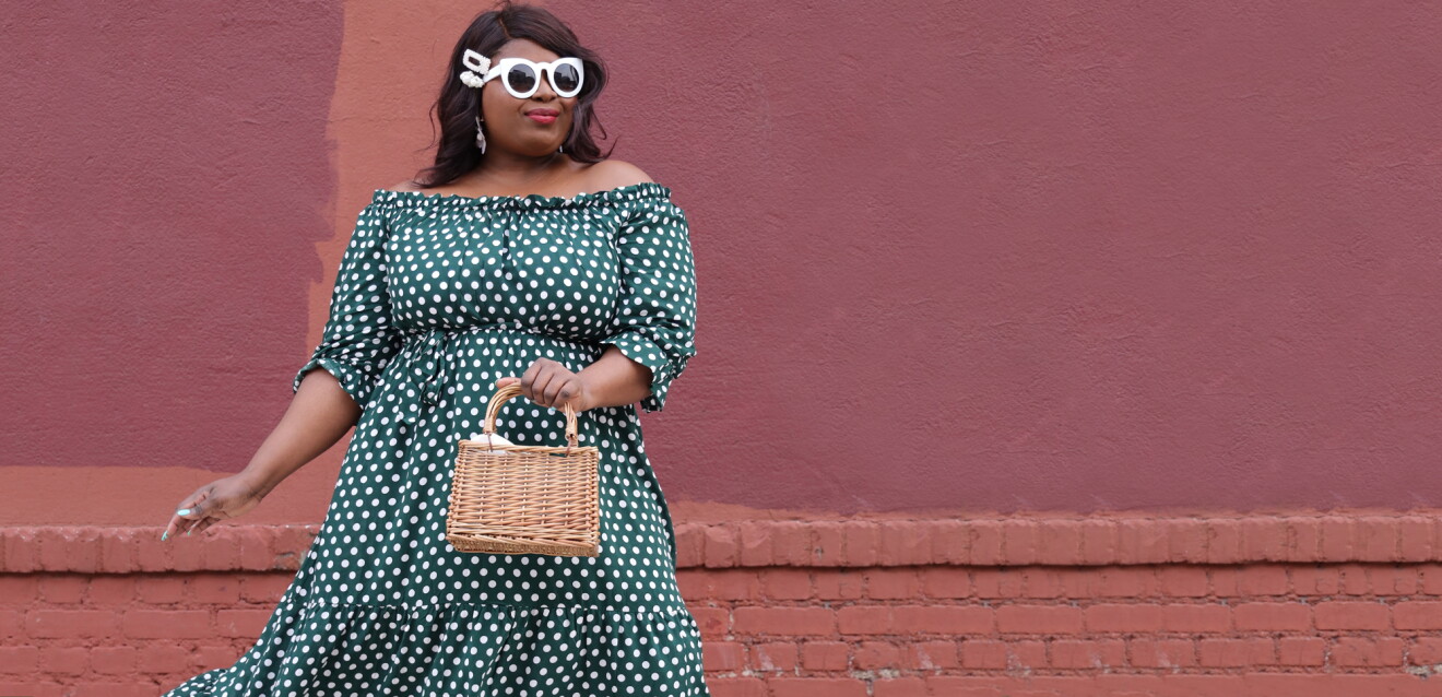 A stylish woman in front of a brick and stucco wall wears an off-the-shoulder dress with white polka dots, white retro sunglasses, and carries a small wicker purse.