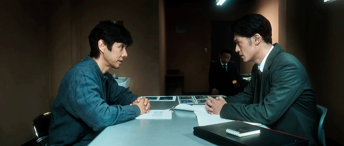 薄暗い取調室で机を挟んで対峙する2人の男性。左側はブルーグレーのシャツ、右側はグレーのスーツを着用。2人とも机の上で両手を組んでいる。机の奥側に複数の写真が並ぶ。奥に立ち会いの男性。