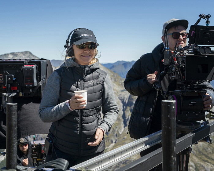 An image of a woman smiling while holding a coffee cup and wearing a headset. She is standing behind a camera directing a shoot on a beautiful hillside.