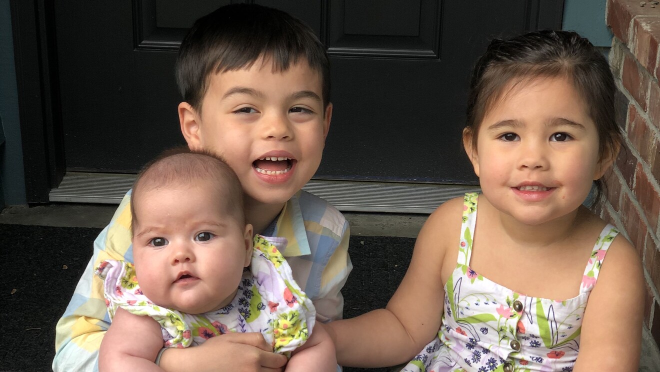 An image of two children smiling for a photo on a doorstep. One child is holding a baby and they appear to be siblings dressed in color-coordinated outfits.