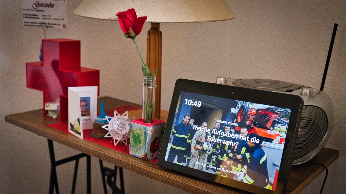 Auf einem kleinen Holztisch steht ein Amazon Echo Show an einen CD-Player angelehnt. Auf dem Tisch steht außerdem noch eine Rose, eine Karte und eine Spardose.