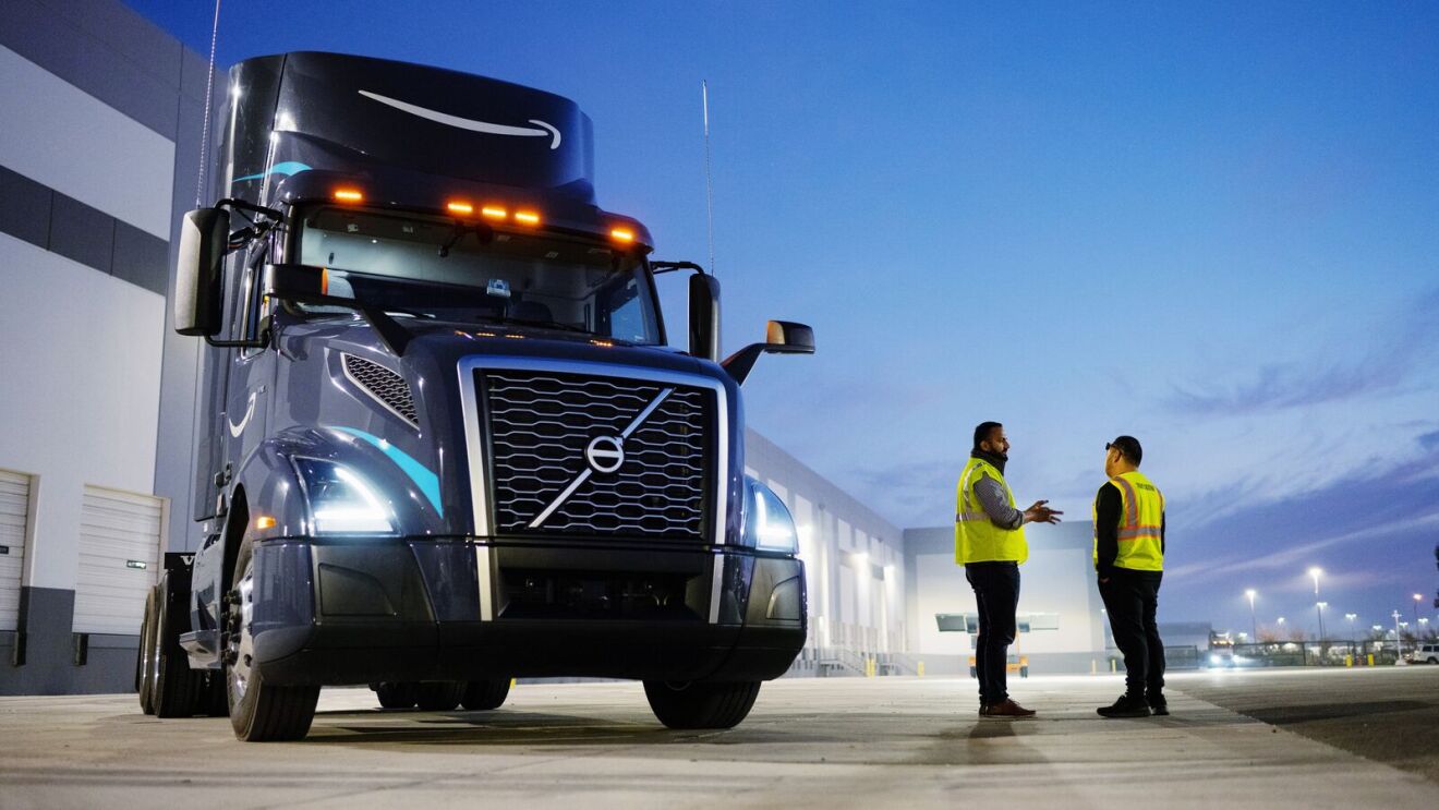 Two men talking outside next to an Amazon delivery truck at dusk.