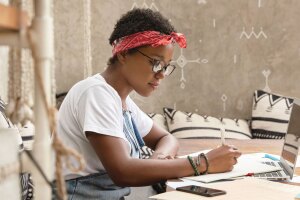 A photo of a person sitting at a desk, writing on a pad of paper that is placed on top of their laptop device.