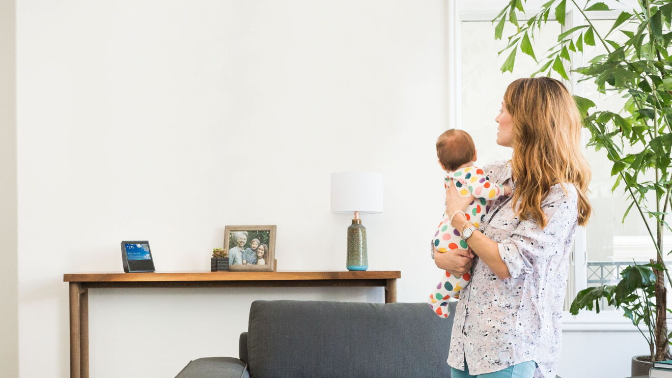 A mom holds her baby as she looks across the living room at a Echo Show device. 