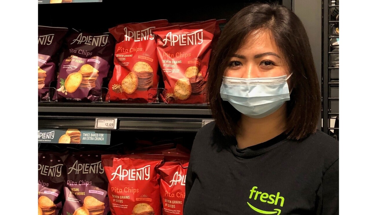 Patricia Payumo wears a Amazon Fresh black T-Shirt as she smiles for the camera standing in front of Aplenty Pita Chips. She is wearing a face mask and has short dark hair.