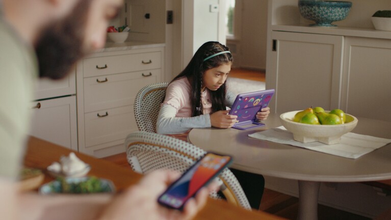 Girl sits at table viewing her tablet while dad is nearby on his smartphone.