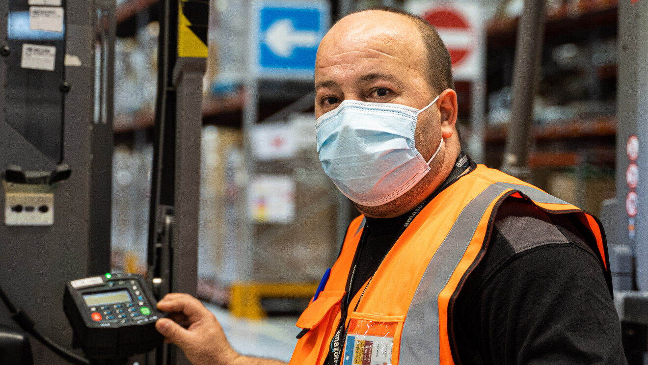 Juan Carlos Moreno, técnico de mantenimiento en el centro logístico de Illescas . Juan Carlos con la mascarilla mirando a cámara con un toro delante. De fondo estanterías llenas de paquetes. Juan Carlos es calvo y el poco pelo que tiene lo tiene corto. Va con un chaleto naranja y lleva el badge colagos. Es moreno y lleva barba de un par de días. 