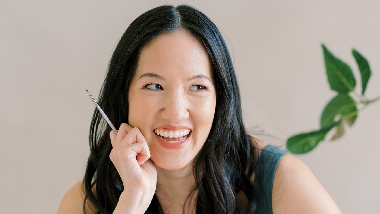 An image of a woman smiling for a photo while leaning her face on her hand and looking away. She is holding a pencil in one hand.