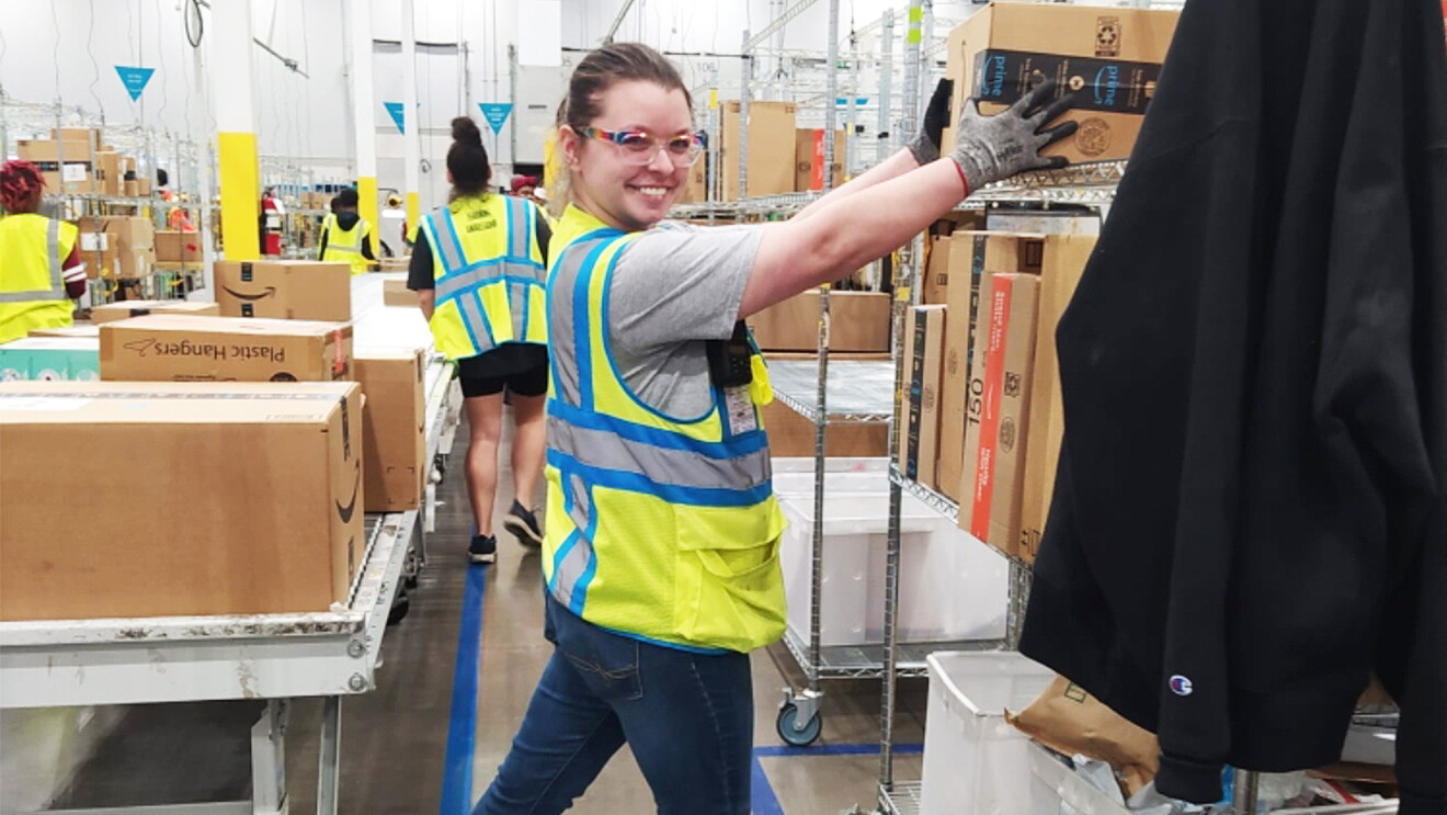 A woman wears a yellow safety vest and glasses as she puts am Amazon box on a shelf in a fulfillment center.