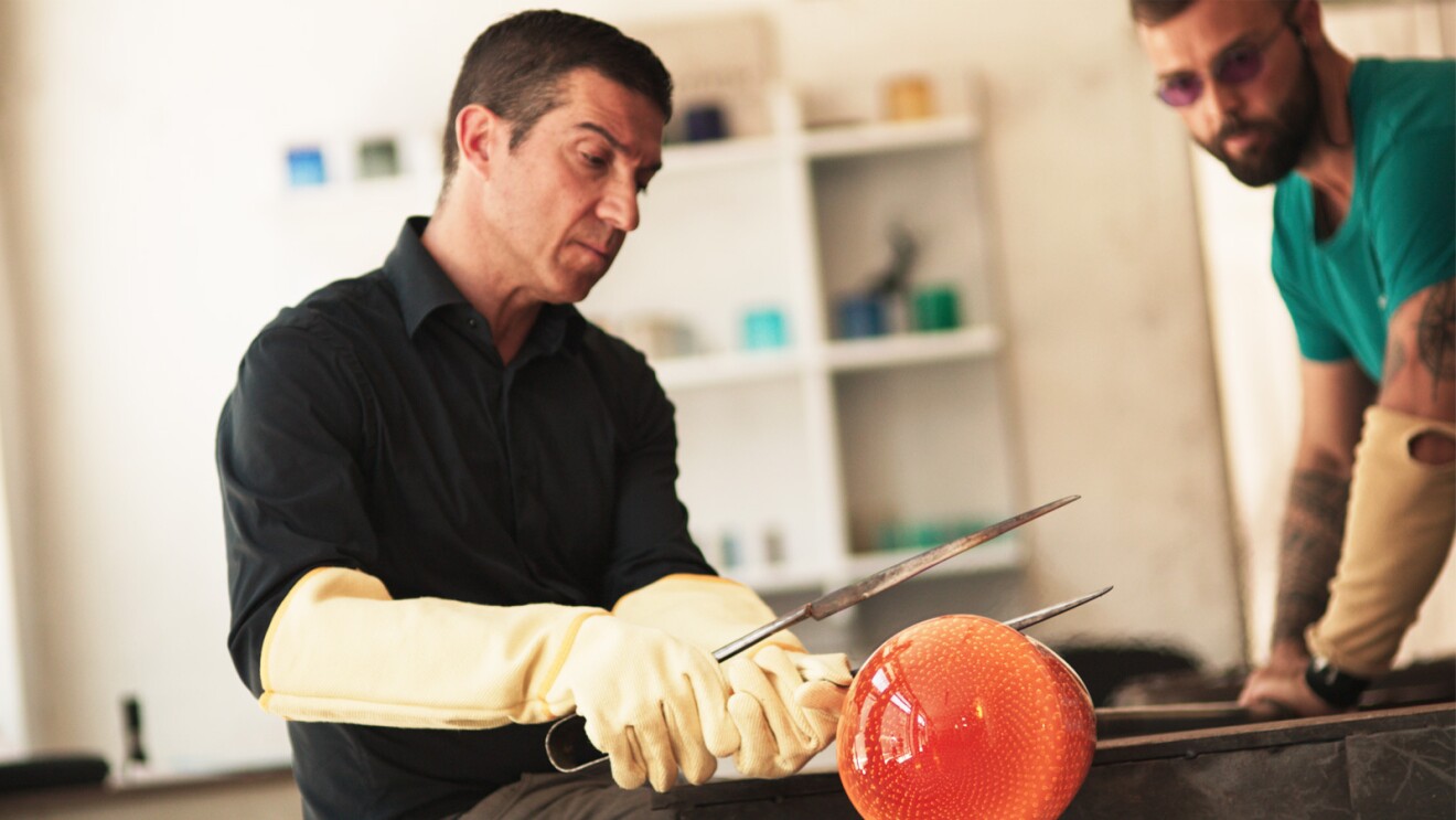 Simone works in his shop blowing and shaping glass. He wears big protective gloves and holds the glass with a big metal tong.