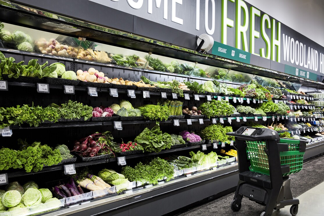 An image of the fresh produce section at an Amazon Fresh store. A Dash Cart sits in front of the aisle and there are green and purple veggies stacked on the shelves.