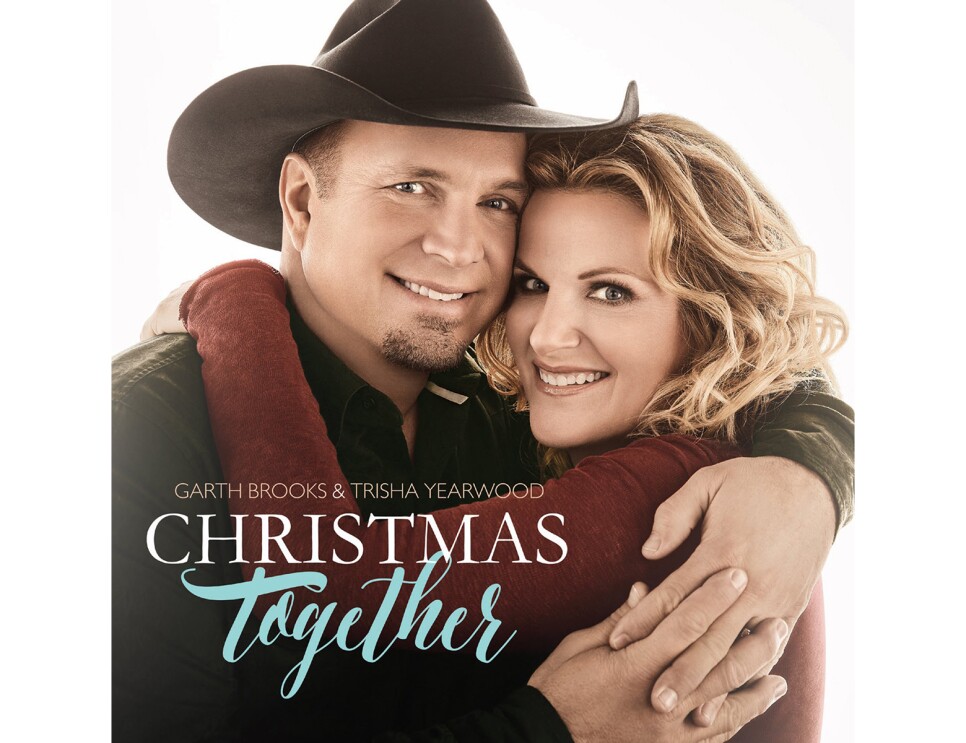 Garth Brooks and Trisha Yearwood face the camera, smiling, as they share an embrace. Garth wears a black cowboy hat and button down. Trisha wears her hair in loose curls and a burgundy long-sleeve top.