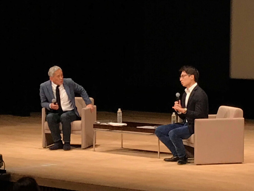 薄暗いホールのステージ上、ソファに腰掛けて対話する2人の男性。2人の間にはローテーブル。