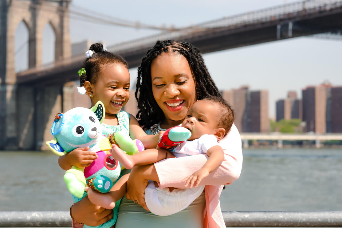 A woman holds a baby and a young child in her arms.  The group is standing in front of a river and a bridge