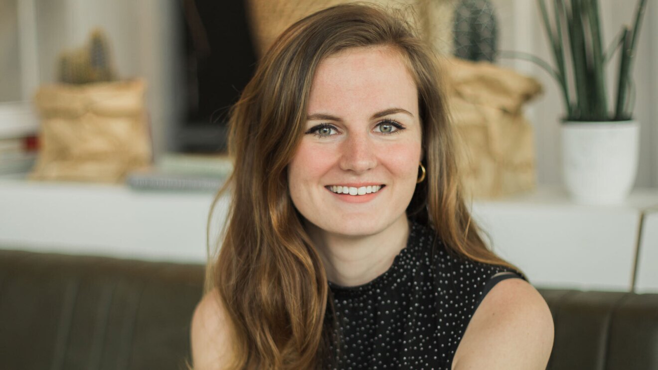 An image of a woman smiling for a headshot photo while sitting on a bench indoors.