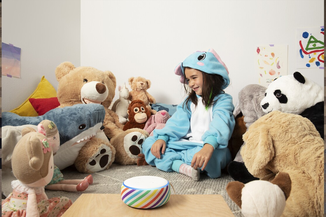 A child sits in a playroom, surrounded by stuffed animals. In front of her is an Echo Dot device is a rainbow stripe, on a tabletop.