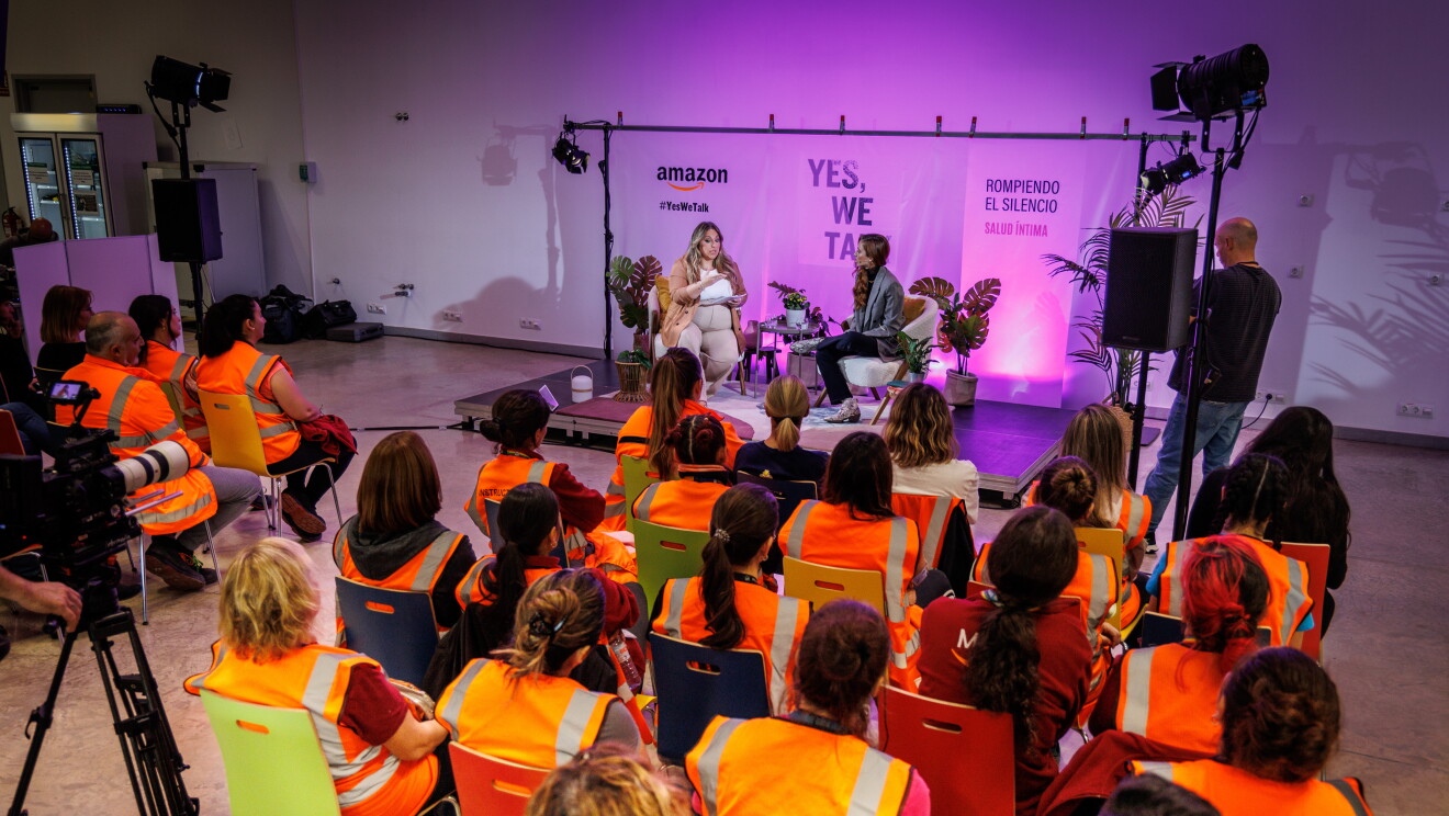 En un escenario creado para la ocasión, de fondo hay un cartel con el logo de Amazon, el Yes, we talk, que es el lema de la campaña y el lema de la sesión Rompiendo el silencio. Todo el fondo es de color rosa. En el escenario, 6 plantes con sus respectivas macetas, dos mesas y dos sillas en la que están sentadas Nicole, una trabajadora de Amazon, y la directora de cine Leticia Dolera. Nicole va vestida a tonos beis. Con un pantalón acampanado, un jersey y una americana. Lleva el pelo largo, ondulado, castaño con mechas rubias, Leticia Dolera va con una americana gris, jersey negro de lana de cuello alto, pantalón negro y botines animal print. Es pelirroja, lleva el pelo largo hasta el pecho y todo a un lado. Las dos están hablando. Ellas aparecen en el fondo, en primer plano trabajadores de Amazon asistiendo a la sesión. La mayoría son mujeres y van con un chaleco de color naranja. 