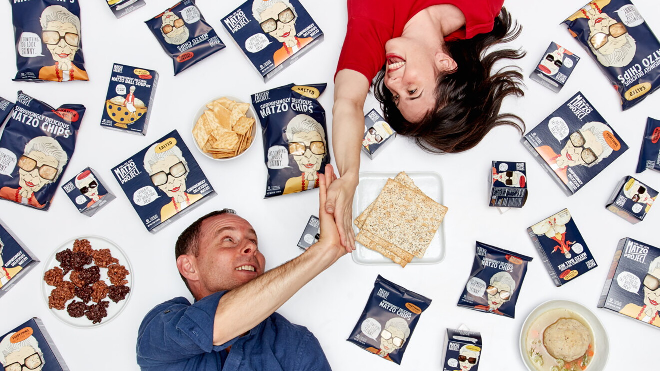 A woman lies on her back, with her right hand extended above her head as she gives a man, also lying on the ground, a high five. Around them are packages of matzo and other products. 