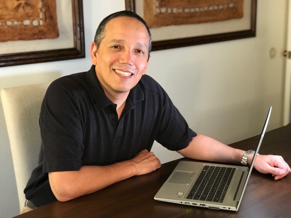 An image of a man smiling for a photo while sitting at a dining table with his laptop in front of him.