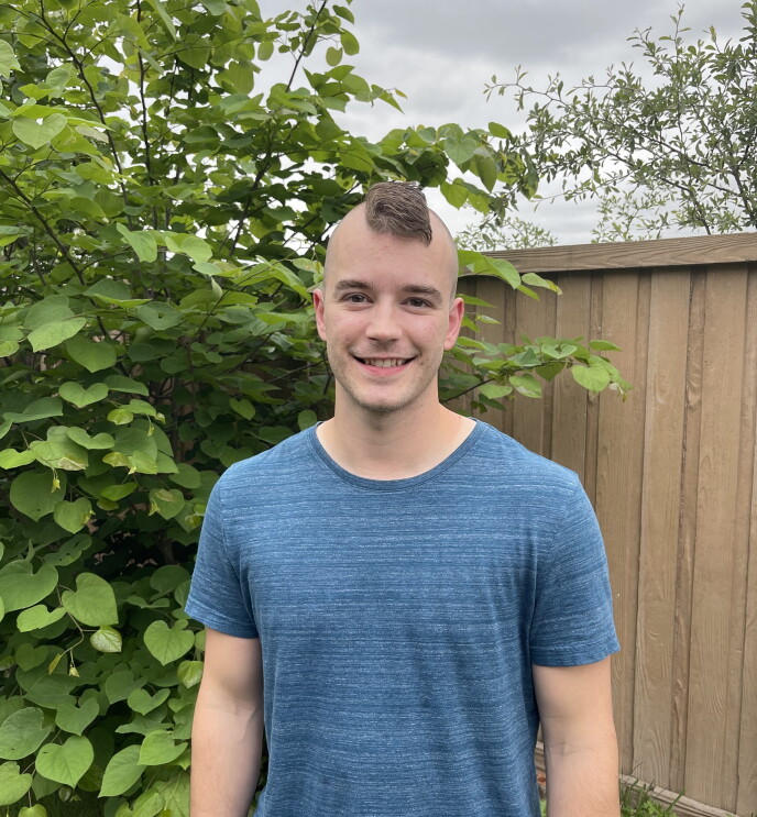 Bryce smiling, wears a blue T-Shirt and has a mohawk. He is standing in front of a bush and a tan fence.
